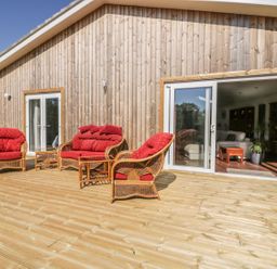 A wooden deck with wicker furniture and red cushions outside a wooden house with glass doors at Chy Lagen Hos in Newquay
