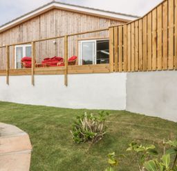 A garden with grass and shrubs outside a wooden house with a balcony and red cushions on chairs at Chy Lagen Hos in Newquay