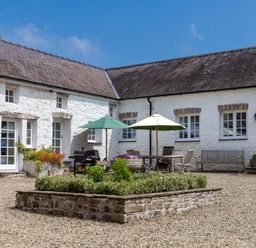 An outdoor courtyard with white stone buildings a raised flower bed a wooden table with umbrellas chairs and a bench at The Old Carriage Court in Kidwelly