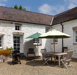 An outdoor patio area with a wooden dining table and chairs under umbrellas and a barbecue grill in front of a white stone building at The Old Carriage Court in Kidwelly