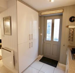 An entrance area with a gray door with glass panels a black doormat white cabinets a kitchen sink and a wall clock with a fish decoration at Serendipity in Fowey