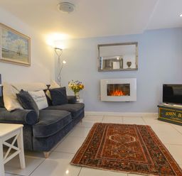 A living room with a dark sofa pillows a side table lamp an electric fireplace a patterned rug and a television at Serendipity in Fowey