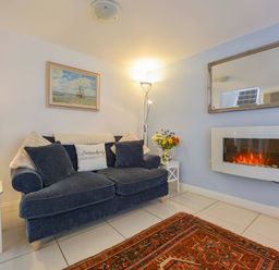 A living room with a blue sofa and cushions a white fireplace a mirror a floor lamp a side table with flowers and a table lamp at Serendipity in Fowey