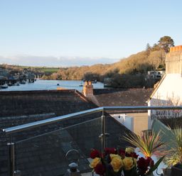 A balcony with flowers overlooking rooftops and a river with boats at The Shell Seekers in Fowey