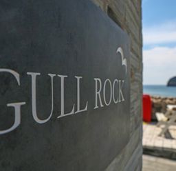 A sign with Gull Rock text on a wall near an outdoor seating area and ocean view at Gull Rock in Trebarwith Strand