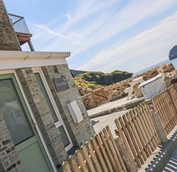 A stone building with a green door and windows next to a wooden fence and a stone wall with a picnic table overlooking the sea at Gull Rock Trebarwith Strand