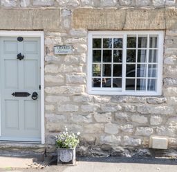 A stone cottage wall with a pale blue door and a window beside it with a flower pot outside at Crooked Cottage in Kirkbymoorside