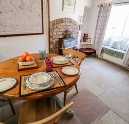 A dining area with a round wooden table set with plates glasses and cutlery a bowl of oranges a brick fireplace and a window seat with cushions at Crooked Cottage in Kirkbymoorside