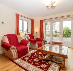 A living room with two armchairs and a coffee table on a patterned rug with glass doors leading outside at Vine Lodge in Kingsbury Episcopi near Martock
