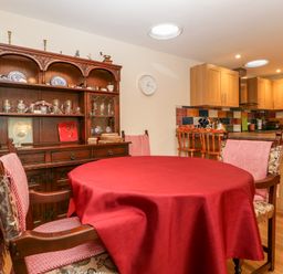A dining area with wooden chairs and a round table covered in a red tablecloth next to a wooden cabinet and kitchen at Vine Lodge in Kingsbury Episcopi near Martock