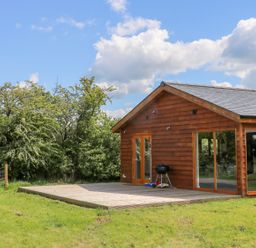A wooden lodge with large windows and a patio with a grill in a grassy area at Oak Lodge in Sheriff Hutton