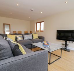 A living room with grey sofas and yellow patterned pillows a coffee table with board games a wall-mounted TV and wooden flooring at Rose Lodge in Sheriff Hutton