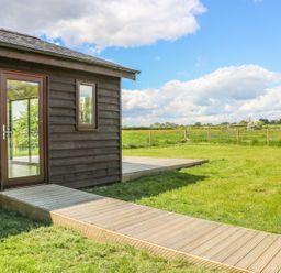 A wooden house with a glass door and window next to a wooden pathway and deck in a grassy field at Rose Lodge in Sheriff Hutton