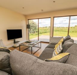 A living room with a grey sofa and patterned cushions a coffee table a television and large windows overlooking a grassy outdoor area at Rose Lodge in Sheriff Hutton