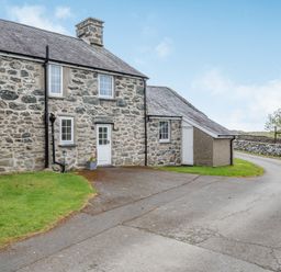 Exterior view of a stone-built house with white doors and windows near a paved driveway and green grass at Gwrtheyrn in Dyffryn Ardudwy
