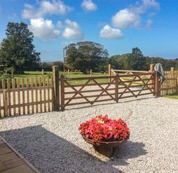 A gravel driveway with a flower pot and wooden gates opening to a grassy field with trees and cows at Edwins View in Nancegollan nr Porthleven