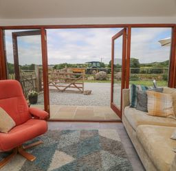 A seating area with a beige sofa and orange armchair next to open glass doors overlooking a garden and field at Edwins View in Nancegollan nr Porthleven