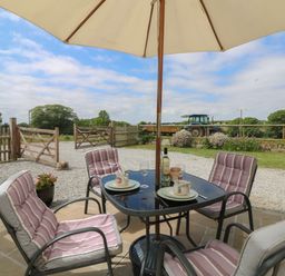 An outdoor patio with a table, four cushioned chairs, an umbrella, and a tractor in the background at Edwin's View in Nancegollan, nr Porthleven