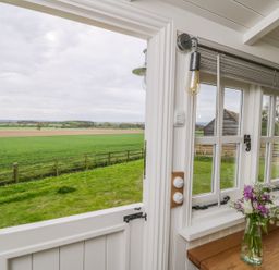 A half door open to a grassy field with a wooden fence outside and flowers with a bottle on a wooden countertop inside at Bellwether in Helmsley