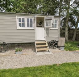 A small gray shepherd hut with wheels stairs and open door outside on a gravel path with a barbecue grill and trees at Bellwether in Helmsley