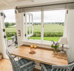 A wooden table with two chairs two glasses of orange juice a plate with croissants a lamp and flowers near an open window and door showing a garden and fields at Bellwether in Helmsley