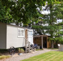 A small wagon-style cabin with stairs leading to an open door beside a wooden shelter with seating and a table with chairs in a garden at Bellwether in Helmsley