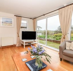 A living room with a wooden floor a gray armchair a coffee table with flowers a television on a white stand and large windows with beige curtains at Wood View Cottage in Blackawton