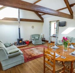 A living room with wooden beams a wood stove two armchairs a sofa a dining table with chairs and flowers at Muncaster View in Carleton near Ravenglass