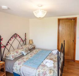 A bedroom with a double bed with floral bedding and blue runner beside a wooden door with a lamp on a wicker table and a vase of flowers on a dresser at Muncaster View in Carleton near Ravenglass