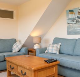 A living room with two blue sofas a wooden coffee table with remote controls a side table with a lamp and a painting of sailboats on the wall at Rose Cottage in Barnstaple