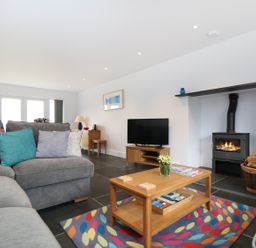 A living room with a grey sofa and colorful cushions a wooden coffee table on a multicolored rug a television on a wooden stand a wood-burning stove with a basket of firewood at Myles View in Pendeen