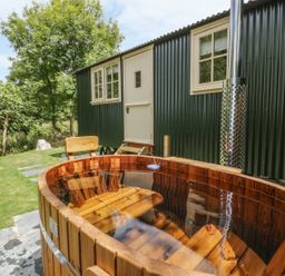 An outdoor wooden hot tub with bench seating next to a green corrugated metal building with white windows and door at Orchard Burlawn near Wadebridge