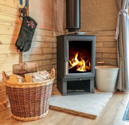 A wood burning stove with fire inside next to a basket of firewood and an ash bucket by a window at Dingle Den in Craswall