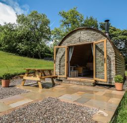 A stone cabin with open wooden doors a wooden picnic table on a paved patio surrounded by grass and trees at Dingle Den in Craswall