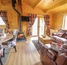 A living room with wooden walls and ceiling a wood burning stove a dining table with chairs and brown leather sofas at Manor Farm Lodges - Red Kite Lodge Newtown