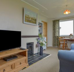 A living room with a TV on a wooden stand a fireplace a vase with flowers a small dining table and chairs and a blue armchair at The Quillet in Pendeen