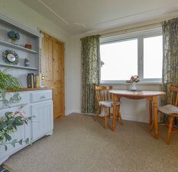 A dining area with a wooden table and two chairs next to a window with curtains and a cabinet with shelves and plants at The Quillet in Pendeen