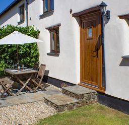An outdoor patio area with a wooden table and chairs under a white umbrella next to a wooden door and windows at Post House in Holsworthy