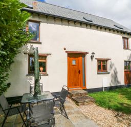An outdoor patio with a glass table and four chairs next to a house with a wooden door and several windows at Post House in Holsworthy