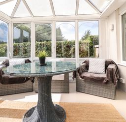 A conservatory with a glass round table and three wicker chairs with cushions at Palm Court in Burton-in-Kendal