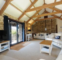 A living room with exposed wooden beams stone walls two sofas a coffee table a television and glass doors at Lily Cottage in Looe