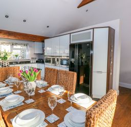 A dining area with a wooden table set with white dishes and glasses next to a kitchen with white cabinets and a black refrigerator at Swn y mor in Amroth