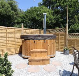 An outdoor area with a wooden hot tub surrounded by a wooden fence and garden chairs at Two Moors Shepherds Hut in Hittisleigh