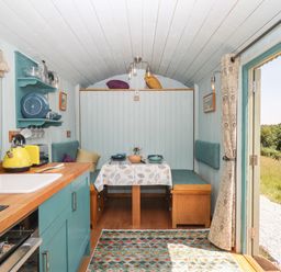 A compact kitchen and dining area inside a small cabin with wood countertops and open doors showing a grassy outside at Two Moors Shepherds Hut in Hittisleigh