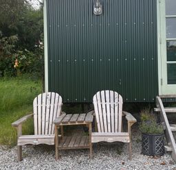 Two wooden chairs and a small table outside a green building at Two Moors Shepherds Hut Hittisleigh