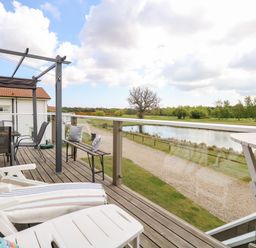 A balcony with lounge chairs a bench and a table overlooking a pond and trees at Links View in Fritton