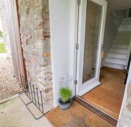 An entrance with a glass door opening to a hallway with white stairs and a doormat outside the door at Whitehall Farm Cottage in Honiton