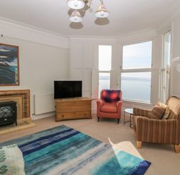 A living room with armchairs a television on a wooden cabinet a fireplace and a large window overlooking the sea at 11 Marine Terrace in Criccieth