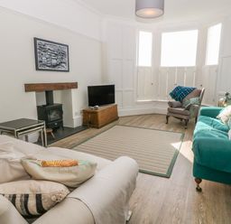 A living room with a beige armchair a green sofa a gray armchair with a throw pillows a framed picture a wood stove and a television at 11 Marine Terrace in Criccieth