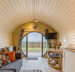 An interior of a wood-paneled pod with a grey sofa with cushions, a round window with curtains, a wall-mounted TV, a small dining table with chairs, and kitchen cabinetry at Corndon Pod White Grit near Chirbury